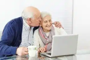 An elderly couple using a laptop and the husband is kissing his wife.